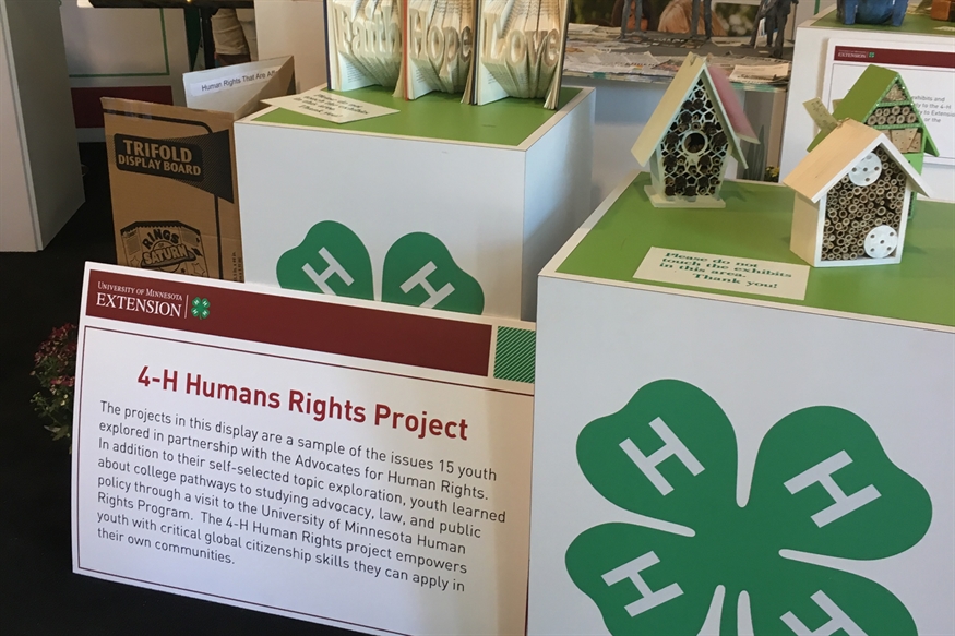 [ai] An exhibit featuring the 4-H Humans Rights Project, showcasing various projects by youth. A sign with red text provides information about the projects, alongside decorative items and a green four-leaf clover logo.